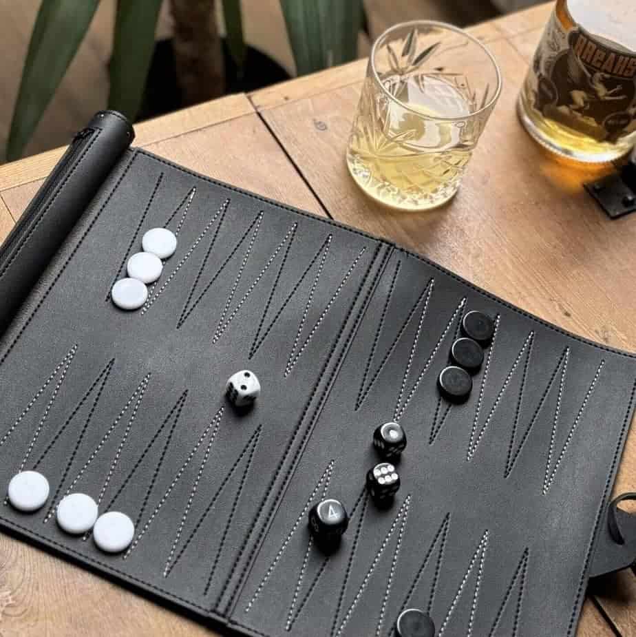 Elegant backgammon-spil i sort læder fra hobbysport.dk, opstillet på et træbord med whiskyglas ved siden af.